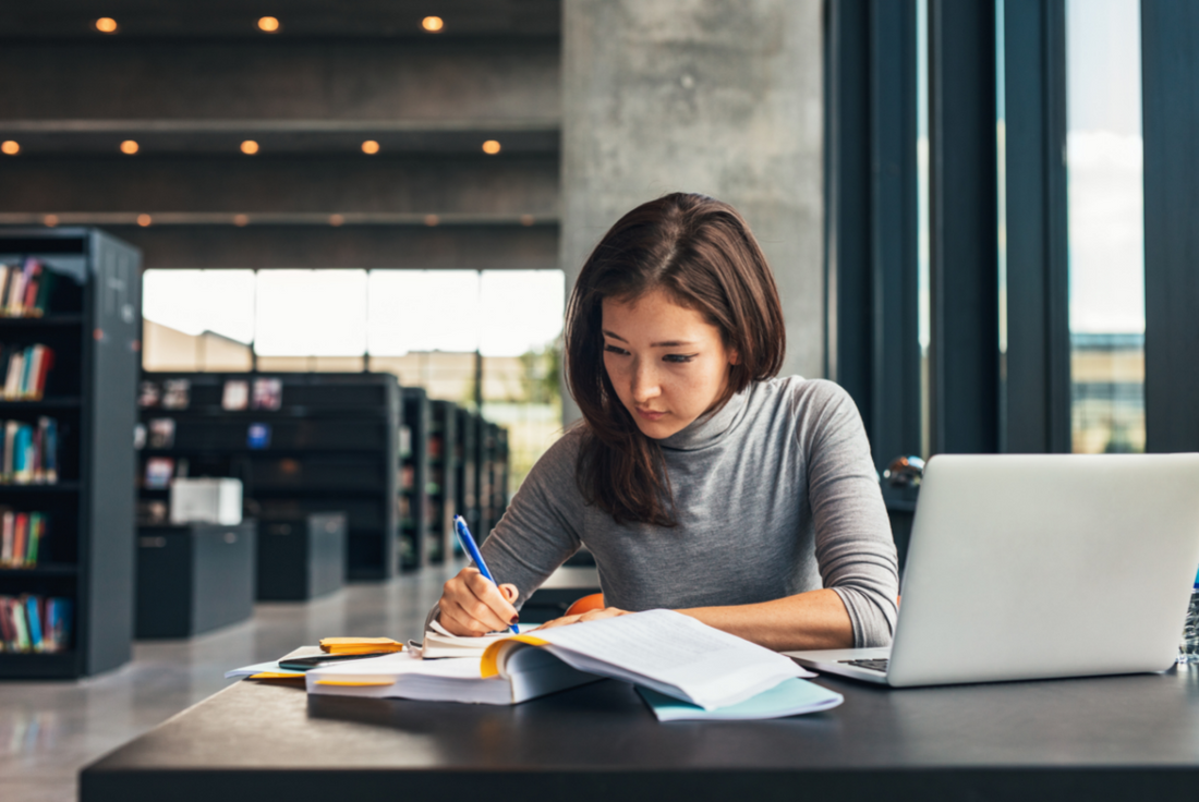 Woman sitting at a table with books and laptop studying for the MCAT exam.